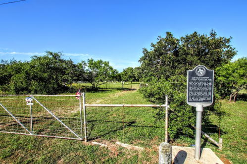 A gated entrance to a grassy area with a historical marker and trees under a clear blue sky.