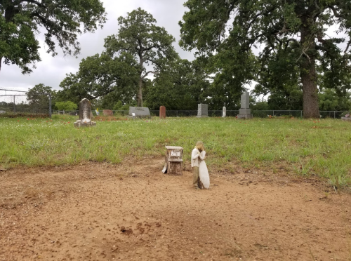 A small angel statue stands on a grassy area in a cemetery, with gravestones visible in the background.