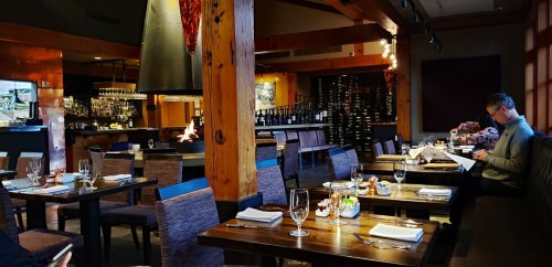 A cozy restaurant interior with wooden beams, tables set for dining, and a man reading a menu by a fireplace.