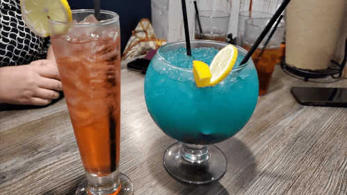 Two colorful cocktails on a wooden table: a tall glass with a pink drink and a large bowl with a blue drink and lemon slices.