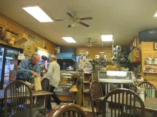 A cozy café interior with wooden walls, tables, and a counter; two people are moving about, one carrying a bag.