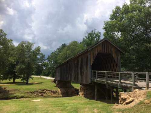 A wooden covered bridge spans a grassy area, surrounded by trees under a cloudy sky.