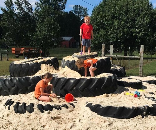 Three children play on a tire structure filled with sand, with trees and a barn in the background.