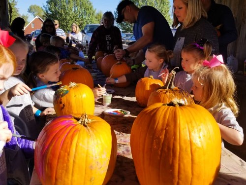 Children painting pumpkins at a table, with adults supervising in a sunny outdoor setting.