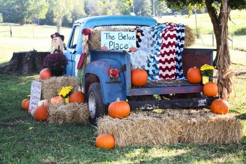 A vintage blue truck decorated with pumpkins, hay bales, and a scarecrow in a sunny outdoor setting.
