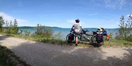 A cyclist stands beside a bike on a gravel path, overlooking a serene lake surrounded by greenery under a blue sky.