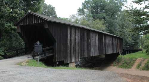 A wooden covered bridge spans a small creek, surrounded by lush greenery and a gravel path leading to it.