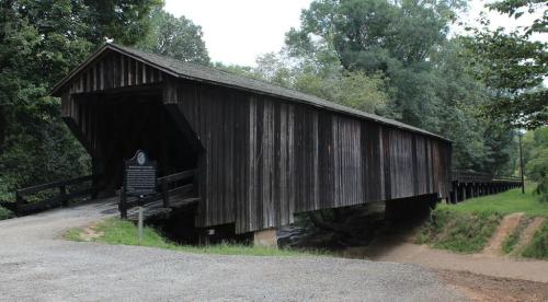 A wooden covered bridge spans a small creek, surrounded by lush greenery and a gravel path leading to it.