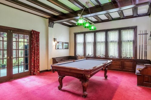 A spacious room with a pool table, green pendant lights, and large windows, featuring wooden beams and red carpet.