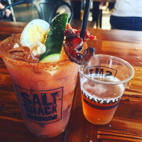 A colorful cocktail garnished with bacon, cucumber, and an egg, alongside a small glass of beer on a wooden table.