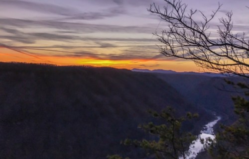 Sunset over a valley with a winding river, framed by silhouetted trees and a colorful sky.