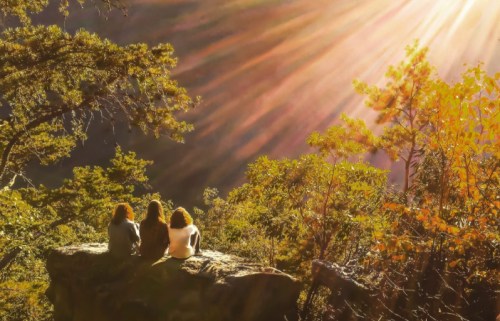 Three people sit on a rock, gazing at a sunlit landscape with rays of light filtering through trees.