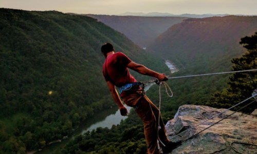 A climber leans back over a cliff, secured by a rope, with a scenic view of a river and mountains in the background.