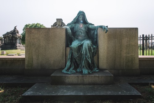 A bronze statue of a seated figure draped in robes, set against a stone backdrop in a cemetery.