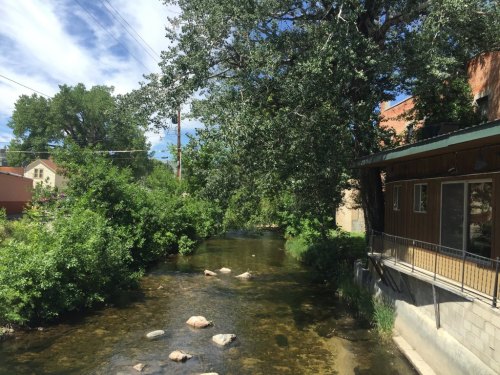 A serene river flows beside a building, surrounded by lush greenery and trees under a blue sky with scattered clouds.