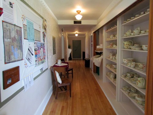 A hallway with wooden floors, a chair, a bulletin board, and shelves displaying various teapots and cups.