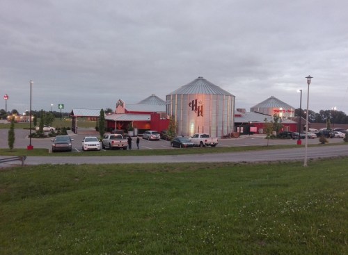A restaurant with a unique silo design, surrounded by a parking lot and green grass under a cloudy sky.