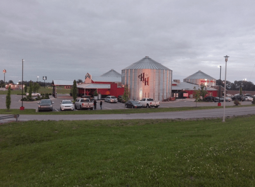 A restaurant with a unique silo design, surrounded by a parking lot and green grass under a cloudy sky.