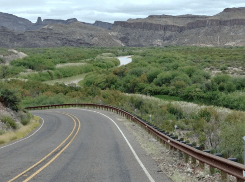 A winding road curves alongside a river, surrounded by lush greenery and rocky hills under a cloudy sky.