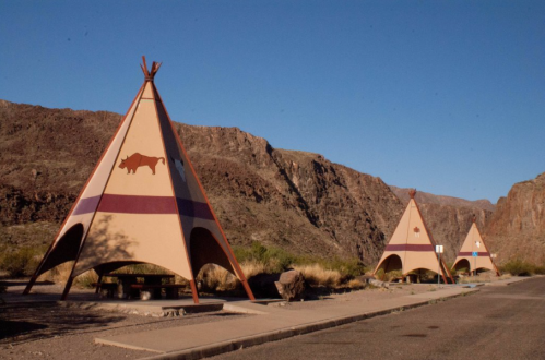 Three colorful teepees stand along a road, set against a backdrop of rocky mountains and clear blue sky.