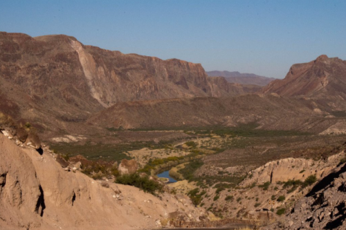 A vast desert landscape with rugged mountains and a winding river below, under a clear blue sky.
