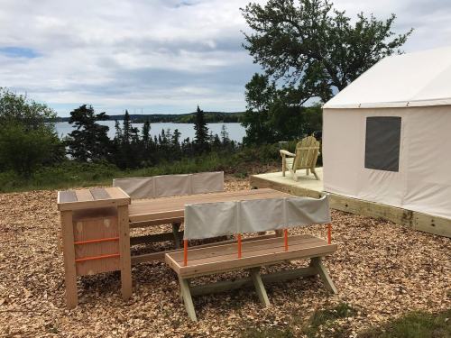 A wooden picnic table and benches near a glamping tent by a lake, surrounded by trees and a cloudy sky.