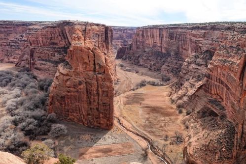 A vast canyon with towering red rock formations and a dry riverbed, surrounded by sparse vegetation under a clear sky.