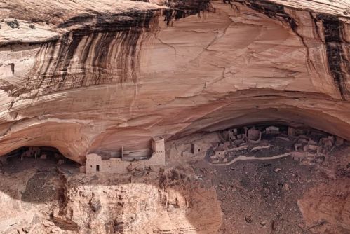 Ancient cliff dwellings nestled under a large rock overhang, surrounded by rugged terrain and desert landscape.
