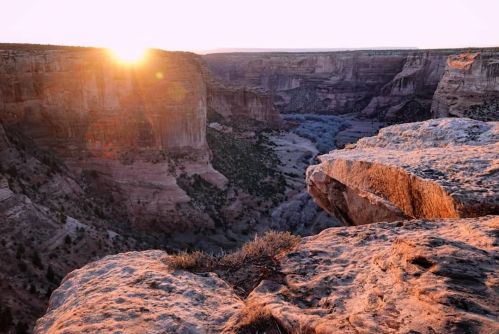 Sunrise over a canyon, casting warm light on rocky cliffs and a winding river below, surrounded by rugged terrain.