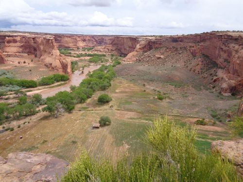 A scenic view of a canyon with a river, green trees, and fields under a cloudy sky.