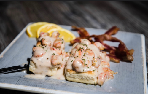 A plate with two biscuits topped with shrimp in creamy sauce, garnished with parsley, and served with bacon and lemon slices.