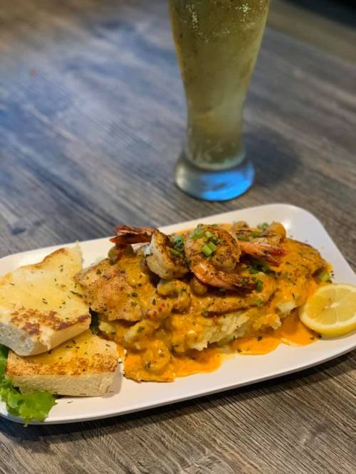 A plate of fish topped with shrimp in a creamy sauce, served with mashed potatoes, garlic bread, and a lemon wedge.