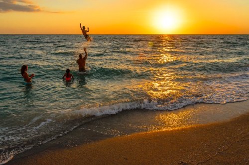 A child is joyfully tossed into the air by an adult in the ocean at sunset, with two others enjoying the water nearby.