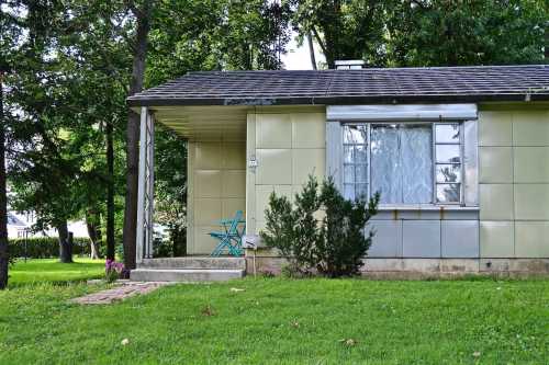 A small house with a green exterior, featuring a front porch and a blue chair, surrounded by trees and grass.