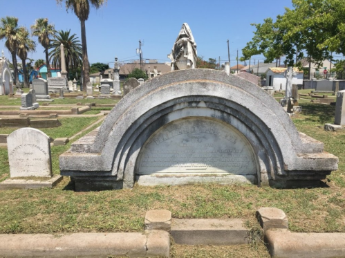 A cemetery scene featuring a large arch-shaped gravestone surrounded by smaller headstones and palm trees.