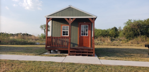 A small green cabin with a red door and porch, set in a grassy area with trees in the background.