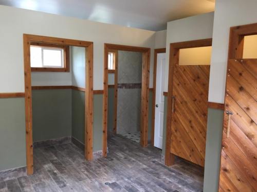 Interior of a bathroom with wooden trim, featuring three stalls and a shower area, all in neutral colors.