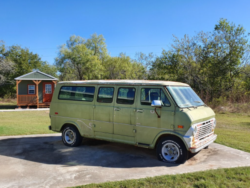A vintage green van parked on a concrete slab, with a small cabin in the background and trees surrounding the area.