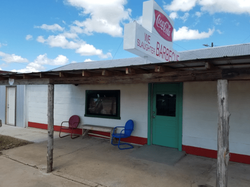 A rustic barbecue restaurant with a Coca-Cola sign, featuring a wooden porch and colorful chairs outside.