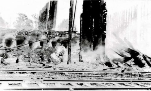Black and white photo of a damaged building with smoke, debris, and railway tracks in the foreground.