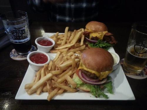 Two burgers with cheese and bacon, served with fries and dipping sauces, alongside two glasses of beer.