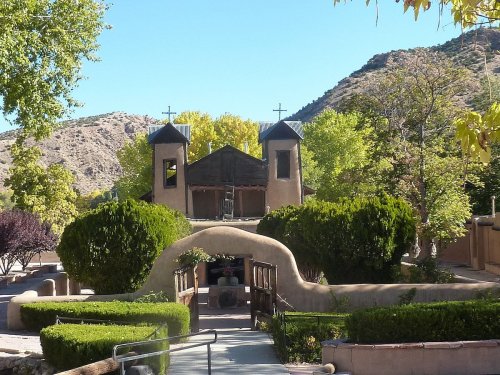 A scenic view of a church with two towers, surrounded by greenery and mountains under a clear blue sky.