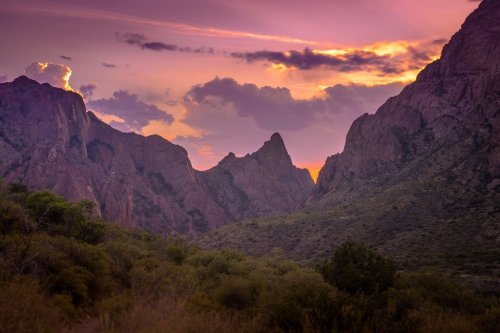 A stunning sunset over rugged mountains, with vibrant purple and orange hues illuminating the sky and landscape.