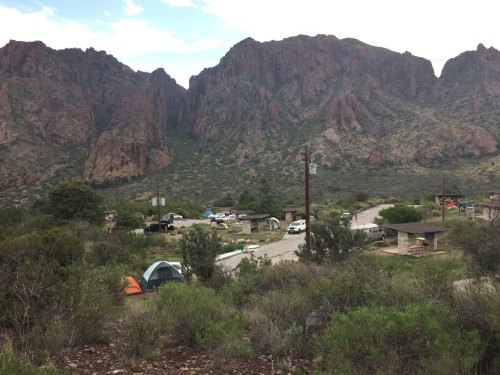 A scenic view of a campsite surrounded by mountains, with tents, vehicles, and picnic areas visible.