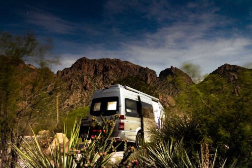 A camper van parked near mountains under a starry night sky, surrounded by desert vegetation.