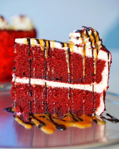 A slice of red velvet cake with cream cheese frosting, drizzled with caramel and chocolate sauce on a silver plate.