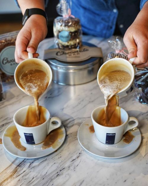 Two cups on a marble surface are being filled with coffee from pouring mugs, with a jar of treats in the background.