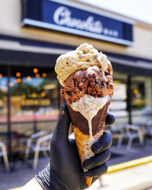 A hand holding a large ice cream cone with chocolate and vanilla flavors, in front of a chocolate shop.