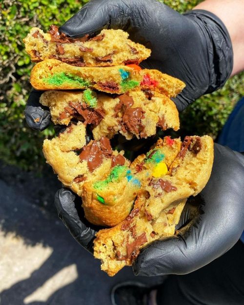 A person wearing black gloves holds a gooey, colorful cookie filled with chocolate and candy pieces.