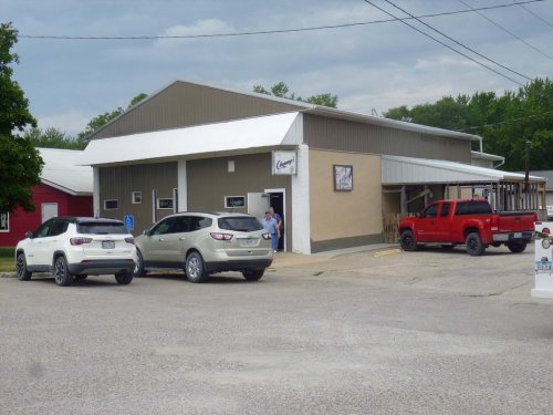 A small commercial building with a metal roof, parked cars in front, and a person standing by the entrance.